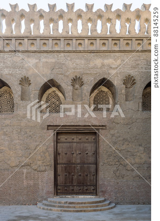 Wooden weathered door, perforated stucco window decorated with floral patterns, and three steps on stone bricks wall 88145259
