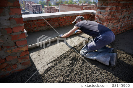 Male laborer in work clothes using screed rail while screeding floor at construction site. Man worker flattening and smoothing surface with straight edge in building with brick walls. Male laborer in work clothes using screed rail while screeding floor at construction site. Man worker flattening and smoothing surface with straight edge in building with brick walls. 88145900