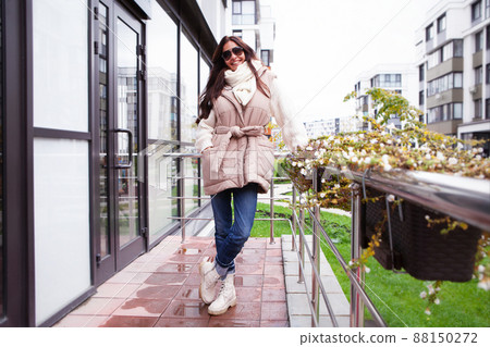Beautiful model wearing beige gilet, warm knitted sweater, jeans pants, high flat leather ankle boots, lace up fastening. Woman smiles on street Beautiful model wearing beige gilet, warm knitted sweater, jeans pants, high flat leather ankle boots, lace up fastening. Woman smiles on street 88150272