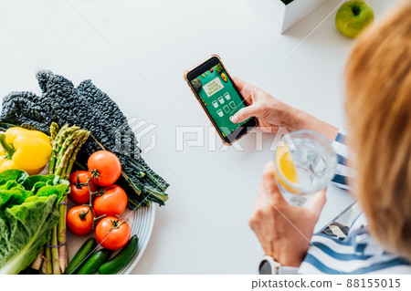 Healthy habit to drink water. Middle age woman drinking glass of pure water with lemon and checking phone while sitting on her kitchen. Using mobile app to control body hydration, Track water balance. 88155015