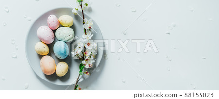 Top view Colored Easter eggs and blooming tree branch on the gray plate with flower petals on the white table. Happy Easter and springtime wide banner. Minimalism. Selective focus. Copy space. Top view Colored Easter eggs and blooming tree branch on the gray plate with flower petals on the white table. Happy Easter and springtime wide banner. Minimalism. Selective focus. Copy space. 88155023