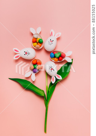 Top view Easter pink background with flower shape made of funny eggs containers with bunny ears filled with candy chocolate eggs. Bunny rabbit symbol. Easter egg hunt. Creative Easter.Vertical card. Top view Easter pink background with flower shape made of funny eggs containers with bunny ears filled with candy chocolate eggs. Bunny rabbit symbol. Easter egg hunt. Creative Easter.Vertical card. 88155025