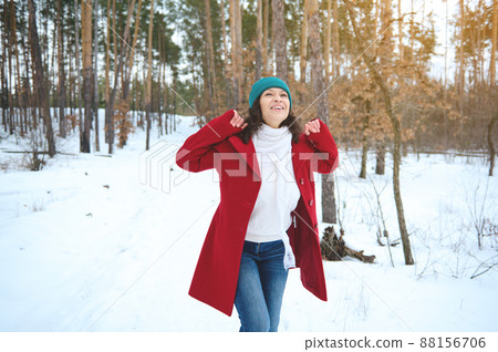 Attractive middle aged woman in bright colorful warm clothes and white woolen scarf whirling around herself, smiles, rejoices, enjoying her weekend in the snow covered forest on a sunny snowy day 88156706