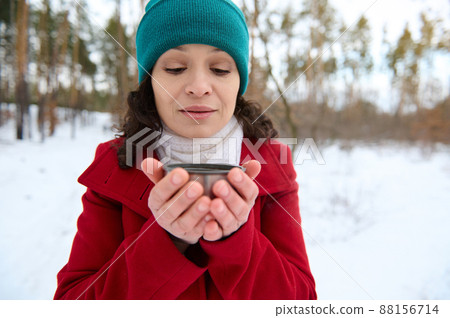 Beautiful middle aged Caucasian woman in green knit hat, red winter coat and white scarf holding a thermo mug from a vacuum flask with hot drink and warming her hands while resting in a snowy forest 88156714