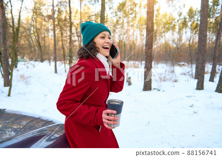 Curly-haired brunette woman driver traveler having fun conversation on mobile phone, smiles cheerfully holding a thermo mug and enjoying a coffee break outdoor sitting on the car hood in snowy forest 88156741