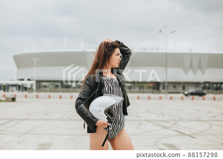 Side view of slim brunette biker girl in bodysuit and leather jacket with safety bike helmet in hands posing at empty stadium outdoors Side view of slim brunette biker girl in bodysuit and leather jacket with safety bike helmet in hands posing at empty stadium outdoors 88157298