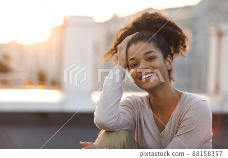 Young happy african american girl looking at camera while sitting on roof terrace on sunny morning Young happy african american girl looking at camera while sitting on roof terrace on sunny morning 88158357