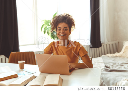 Cheerful young Afro American female student studying online, sitting at desk surrounded by textbooks Cheerful young Afro American female student studying online, sitting at desk surrounded by textbooks 88158358