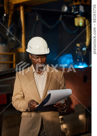Serious foreman in work helmet reading manufacturing plan while controlling the work in factory 88163756