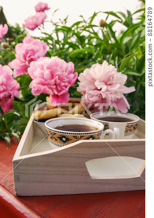 Tea in country style in summer garden in the village. Two cups of black tea on wooden tray and blooming peony flowers in sunlight. Tea in country style in summer garden in the village. Two cups of black tea on wooden tray and blooming peony flowers in sunlight. 88164789