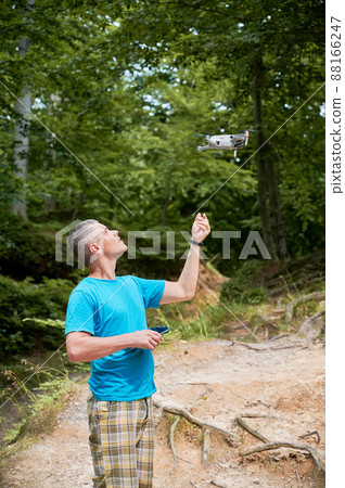 Man operating drone using remote controller. Man using drone for photos and video making while standing in green forest. Man operating drone using remote controller. Man using drone for photos and video making while standing in green forest. 88166247