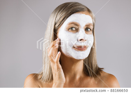 Mature woman with white clay mask on face posing in studio Mature woman with white clay mask on face posing in studio 88166650