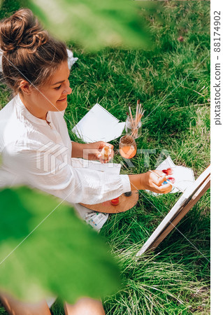 Artist painting on the easel outdoors in the garden. Open air outdoor art workshop. Draw on the canvas with brush and palette sitting on the grass during a picnic in a park Artist painting on the easel outdoors in the garden. Open air outdoor art workshop. Draw on the canvas with brush and palette sitting on the grass during a picnic in a park 88174902