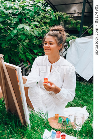 Artist painting on the easel outdoors in the garden. Open air outdoor art workshop. Draw on the canvas with brush and palette sitting on the grass during a picnic in a park Artist painting on the easel outdoors in the garden. Open air outdoor art workshop. Draw on the canvas with brush and palette sitting on the grass during a picnic in a park 88174905