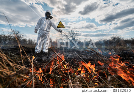 Firefighter ecologist working in field with wildfire. Man in protective suit and gas mask near burning grass with smoke, holding warning sign with skull and crossbones. Natural disaster concept. Firefighter ecologist working in field with wildfire. Man in protective suit and gas mask near burning grass with smoke, holding warning sign with skull and crossbones. Natural disaster concept. 88182920