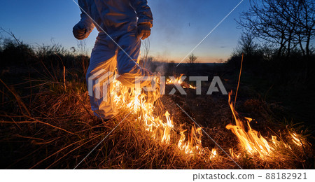 Firefighter ecologist extinguishing fire in field at night. Cropped view of man in protective radiation suit running through burning grass with smoke. Natural disaster concept. Firefighter ecologist extinguishing fire in field at night. Cropped view of man in protective radiation suit running through burning grass with smoke. Natural disaster concept. 88182921