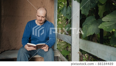Man is reading a paper book sitting on the porch of a village house. The concept of reading paper books, learning and hobbies. Old, retro decor, burlap background 88184489