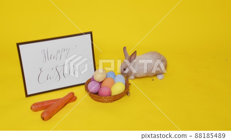 A beige rabbit jumps on a yellow background and sniffs carrots. Next to it is a basket with colorful eggs and a frame with the inscription Happy Easter. Easter. A beige rabbit jumps on a yellow background and sniffs carrots. Next to it is a basket with colorful eggs and a frame with the inscription Happy Easter. Easter. 88185489