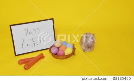 A beige rabbit jumps on a yellow background and sniffs carrots. Next to it is a basket with colorful eggs and a frame with the inscription Happy Easter. Easter. A beige rabbit jumps on a yellow background and sniffs carrots. Next to it is a basket with colorful eggs and a frame with the inscription Happy Easter. Easter. 88185490