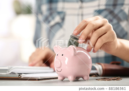 Woman putting money into piggy bank at table indoors, closeup 88186361