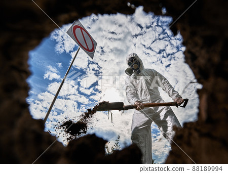 View inside pit of scientist in white suit and gas mask standing near prohibition sign and digging ground to obtain samples and study ground cover in contaminated area. Cloudy sky on the background. View inside pit of scientist in white suit and gas mask standing near prohibition sign and digging ground to obtain samples and study ground cover in contaminated area. Cloudy sky on the background. 88189994
