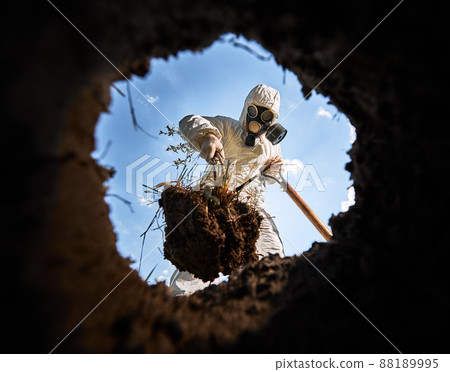 View from inside pit of ecologist digging pit by shovel and planting tree, wearing gas mask and protective overalls outdoors, on background of blue cloudy sky. View from inside pit of ecologist digging pit by shovel and planting tree, wearing gas mask and protective overalls outdoors, on background of blue cloudy sky. 88189995