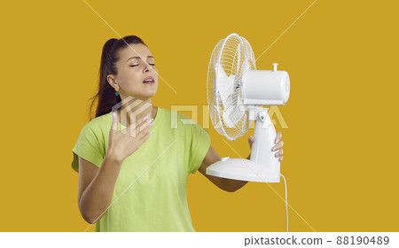 Exhausted young lady having headache because of summer heat, holding fan in her hand is exhausted from the heat, touching her forehead, yellow studio background, need for air conditioner 88190489