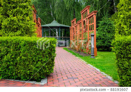 Vertical pergola made of brown wood in a roses garden with a stone tile walkway with granite curb and ground lantern in backyard in a garden with plants and thuja hedge with iron gazebo. Vertical pergola made of brown wood in a roses garden with a stone tile walkway with granite curb and ground lantern in backyard in a garden with plants and thuja hedge with iron gazebo. 88191607