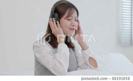 While sitting on her bed at home, a happy young Asian woman in earphones sings and listens to music. Girl dances while listening to music through headphones. A content woman spends her evenings in While sitting on her bed at home, a happy young Asian woman in earphones sings and listens to music. Girl dances while listening to music through headphones. A content woman spends her evenings in 88191658