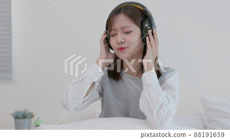 While sitting on her bed at home, a happy young Asian woman in earphones sings and listens to music. Girl dances while listening to music through headphones. A content woman spends her evenings in 88191659