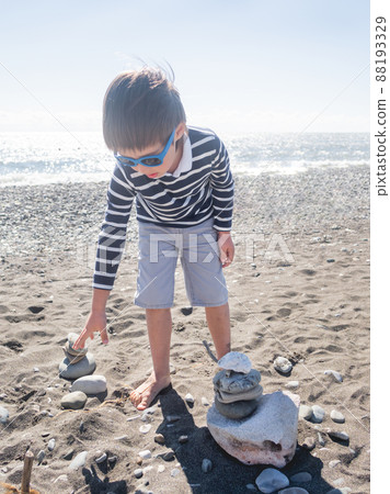 Little boy is building pyramid with stones and driftwood on pebble beach. Vacation on seaside. Exploring nature in childhood. 88193329