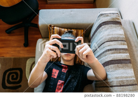 Digital Addiction in Tweens and Teens, smartphone addicted. Teenage boy playing online game, watching messengers in the smartphone laying on the couch at home Digital Addiction in Tweens and Teens, smartphone addicted. Teenage boy playing online game, watching messengers in the smartphone laying on the couch at home 88198280