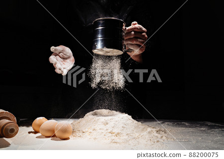 woman sifts flour through sieve onto table with eggs on black background.  88200075