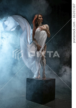 Red-haired girl covers herself with white cloth like an antique statue, posing in the studio 88201387