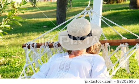 female age 50-60 in a straw hat is resting in a hammock around the palm trees and enjoying the 88202690