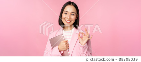 Portrait of corporate woman, girl in office in business suit, holding digital tablet, showing okay, recommending company, standing over pink background Portrait of corporate woman, girl in office in business suit, holding digital tablet, showing okay, recommending company, standing over pink background 88208039