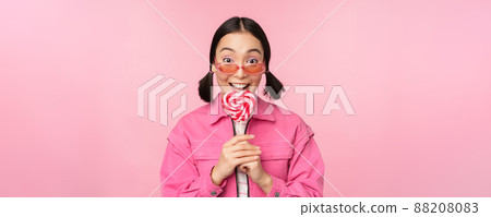 Silly and cute asian female model licking lolipop, eating candy sweet and smiling, looking excited, standing over pink background Silly and cute asian female model licking lolipop, eating candy sweet and smiling, looking excited, standing over pink background 88208083
