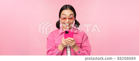Portrait of asian girl in sunglasses using smartphone. Woman looking at mobile phone, browsing in app, standing over pink background Portrait of asian girl in sunglasses using smartphone. Woman looking at mobile phone, browsing in app, standing over pink background 88208084