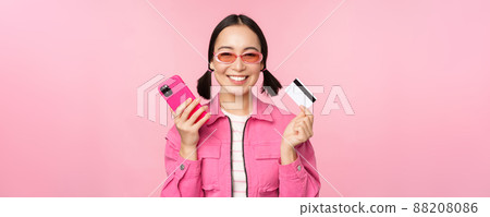 Beautiful korean woman holding smartphone, credit card, smiling at camera, buying online, shopping with mobile phone, standing over pink background Beautiful korean woman holding smartphone, credit card, smiling at camera, buying online, shopping with mobile phone, standing over pink background 88208086