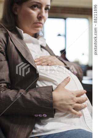 Close up of pregnant woman touching pregnancy tummy while in agency office workspace. Expectant mother sitting in marketing company workplace while holding baby belly bump. Close up of pregnant woman touching pregnancy tummy while in agency office workspace. Expectant mother sitting in marketing company workplace while holding baby belly bump. 88211896