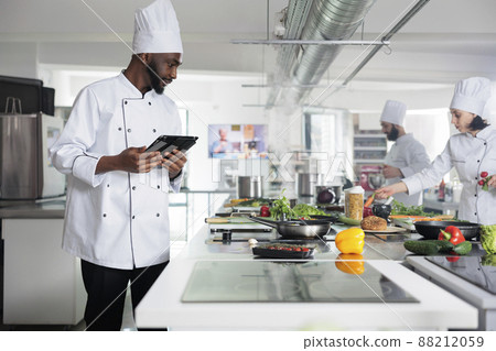 Head cook with tablet brainstorming garnish ideas for gourmet cuisine meal. Food industry worker with handheld touchscreen device preparing ingredients for evening dinner service at restaurant. Head cook with tablet brainstorming garnish ideas for gourmet cuisine meal. Food industry worker with handheld touchscreen device preparing ingredients for evening dinner service at restaurant. 88212059