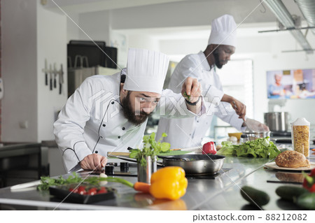 Head chef adding fresh chopped green herbs in pan while cooking gourmet dish in restaurant professional kitchen. Food industry workers preparing delicious meal using organic vegetables. Head chef adding fresh chopped green herbs in pan while cooking gourmet dish in restaurant professional kitchen. Food industry workers preparing delicious meal using organic vegetables. 88212082