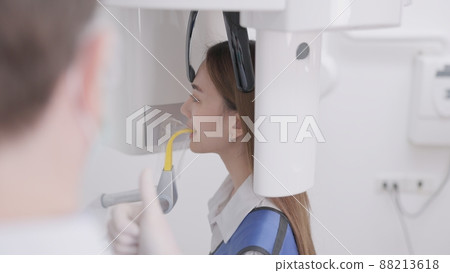 On a panoramic x-ray machine, the dentist aligns the bite of a woman. In a dental clinic, a woman dressed in a lead apron stands still as a panoramic X-ray machine circles her head.. On a panoramic x-ray machine, the dentist aligns the bite of a woman. In a dental clinic, a woman dressed in a lead apron stands still as a panoramic X-ray machine circles her head.. 88213618