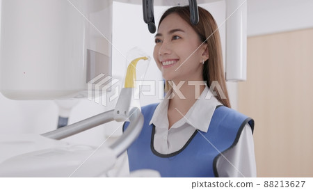 On a panoramic x-ray machine, the dentist aligns the bite of a woman. In a dental clinic, a woman dressed in a lead apron stands still as a panoramic X-ray machine circles her head.. On a panoramic x-ray machine, the dentist aligns the bite of a woman. In a dental clinic, a woman dressed in a lead apron stands still as a panoramic X-ray machine circles her head.. 88213627