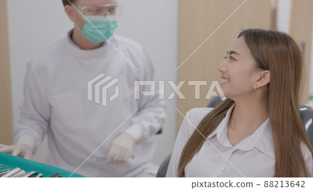 At a sophisticated dental facility, a Caucasian male dentist with a face mask explains how to treat teeth to a cheerful Asian woman patient. At a sophisticated dental facility, a Caucasian male dentist with a face mask explains how to treat teeth to a cheerful Asian woman patient. 88213642