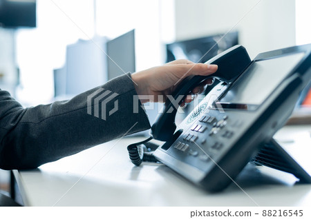 Closeup female hand on landline phone in office. Faceless woman in a suit works as a receptionist answering the phone to customer calls. 88216545