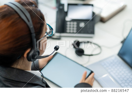 Rear view of a woman in a headset using a digital tablet while sitting at a desk. Friendly female support service operator at work. 88216547