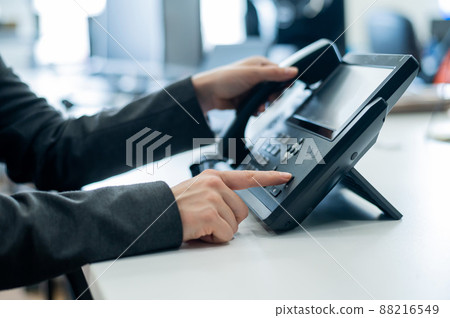 Closeup female hand on landline phone in office. Faceless woman in a suit works as a receptionist answering the phone to customer calls. 88216549