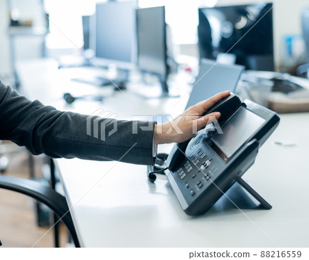 Closeup female hand on landline phone in office. Faceless woman in a suit works as a receptionist answering the phone to customer calls. 88216559