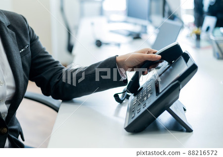 Closeup female hand on landline phone in office. Faceless woman in a suit works as a receptionist answering the phone to customer calls. 88216572